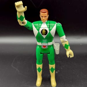 May include: A green action figure of a Power Ranger with a white diamond pattern on the chest and shoulders. The figure has a red belt buckle, a holster, and yellow boots. The figure's hair is reddish-brown, and it is posed with one arm raised.