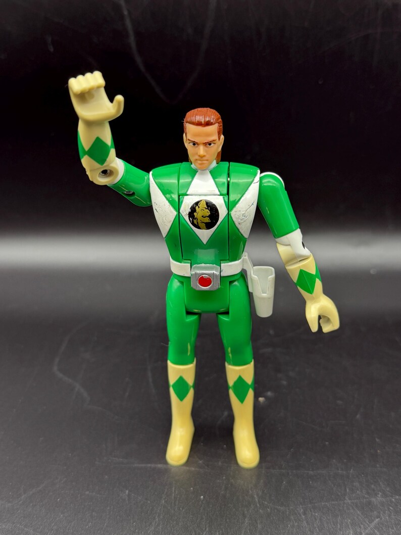 May include: A green action figure of a Power Ranger with a white diamond pattern on the chest and shoulders. The figure has a red belt buckle, a holster, and yellow boots. The figure's hair is reddish-brown, and it is posed with one arm raised.