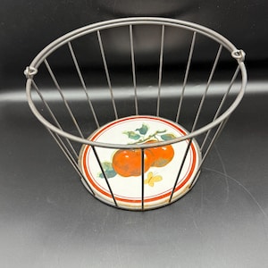 May include: A black metal wire basket with a round, white ceramic base featuring a red apple design. The base has a red and white border.