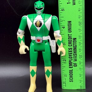 May include: Green Power Ranger action figure with white accents and a gold emblem on the chest. The figure has articulated joints and is wearing a white belt with a holster. A green ruler is next to the figure.