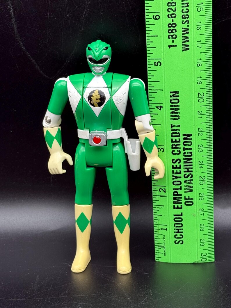 May include: Green Power Ranger action figure with white accents and a gold emblem on the chest. The figure has articulated joints and is wearing a white belt with a holster. A green ruler is next to the figure.