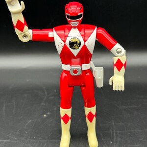 May include: A red Power Ranger action figure with white accents and a gold emblem on the chest. The figure has articulated arms and legs, with white gloves and boots. The figure is a vintage toy.