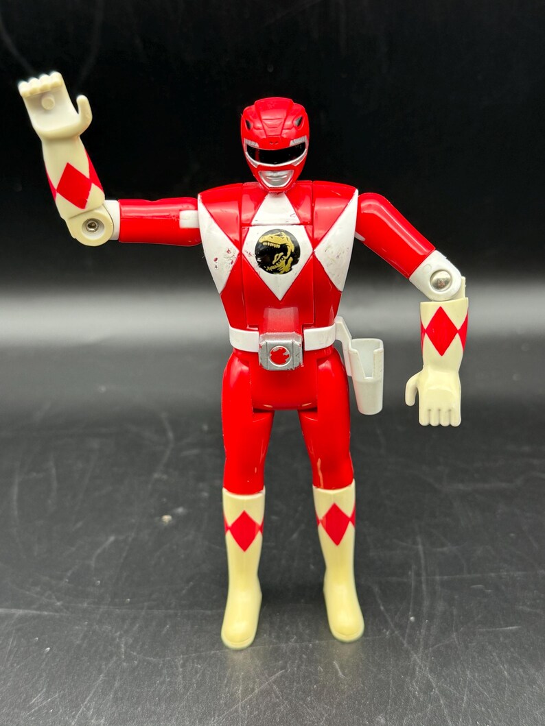 May include: A red Power Ranger action figure with white accents and a gold emblem on the chest. The figure has articulated arms and legs, with white gloves and boots. The figure is a vintage toy.