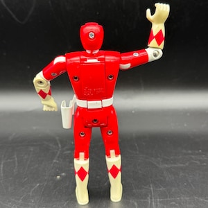 May include: A red action figure with white and cream-colored accents. The figure has a white belt, cream-colored gloves and boots with red diamond patterns. The figure is posed with one arm raised. A white holster is attached to the belt.