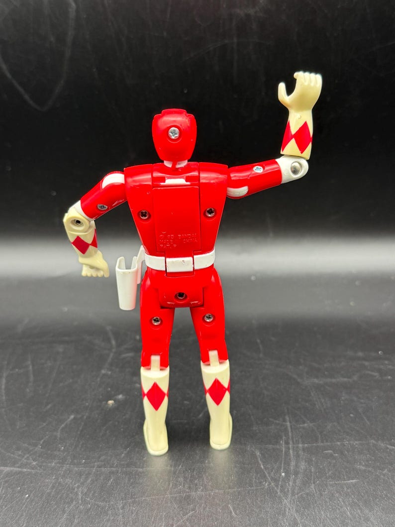 May include: A red action figure with white and cream-colored accents. The figure has a white belt, cream-colored gloves and boots with red diamond patterns. The figure is posed with one arm raised. A white holster is attached to the belt.