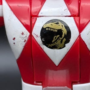 May include: Close-up of a red action figure's chest. White and red geometric shapes frame a black circle with a gold dinosaur emblem. The figure has white accents and visible wear.