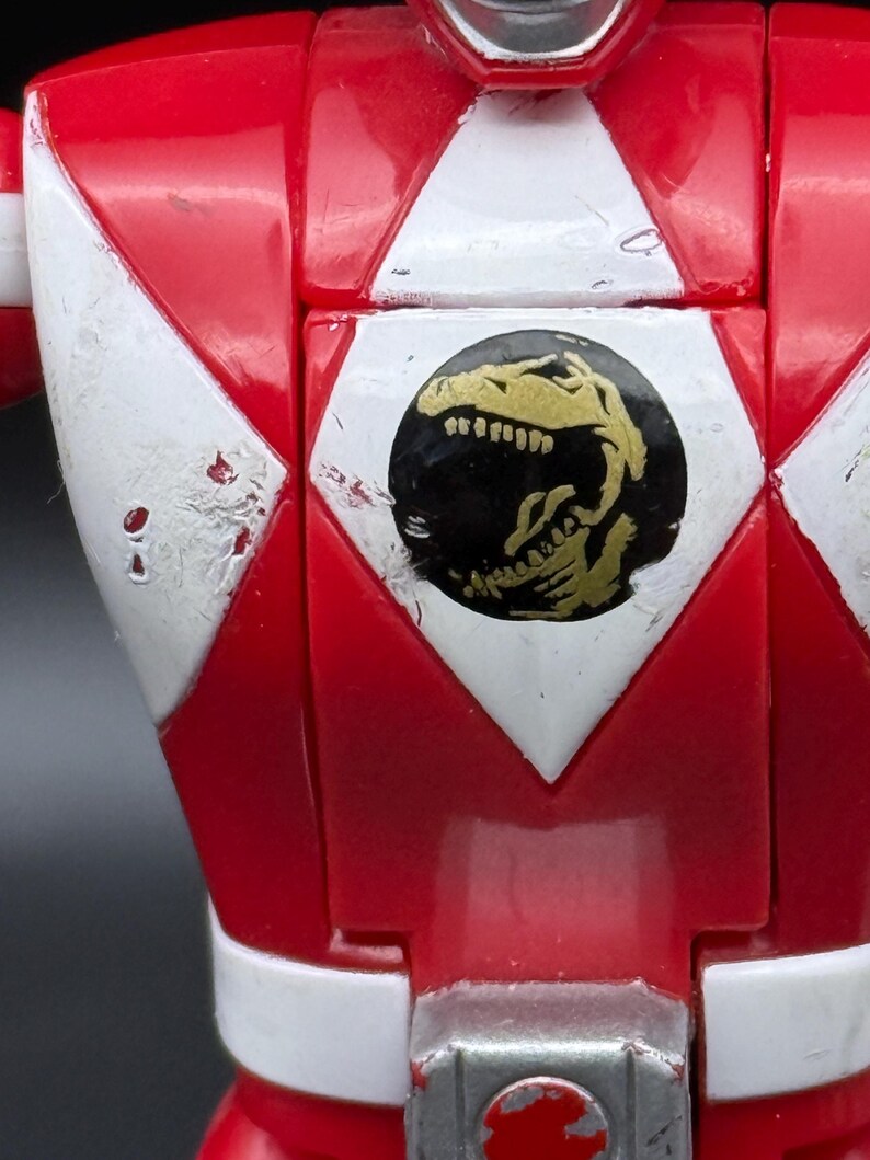May include: Close-up of a red action figure's chest. White and red geometric shapes frame a black circle with a gold dinosaur emblem. The figure has white accents and visible wear.
