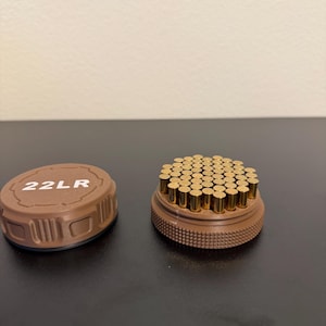 Op de afbeelding: Een bruine container met een deksel met de markering "22LR" naast een container gevuld met talrijke kleine, goudkleurige cilindrische objecten. De containers staan op een donkere ondergrond.