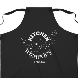 May include: Black kitchen apron with white text that reads "Kitchen Wizardry In Progress" surrounded by white stars.