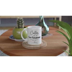 May include: White ceramic coffee mug with the text "Coffee is my love language. Everything else is just small talk." The mug is on a wooden coaster. A small cactus in a green pot and a decorative vase are in the background.