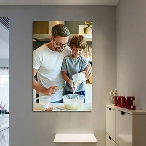 May include: A framed print shows a father and child baking together in a kitchen. The father, wearing a white t-shirt and glasses, assists the child, who is pouring ingredients into a glass bowl. The scene is filled with warmth and the joy of shared activity.