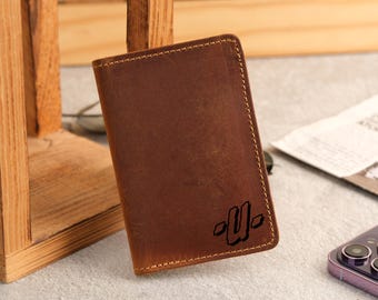 May include: A brown leather wallet with a stitched border and a debossed design in the lower right corner. The wallet is rectangular and appears to be new. The background includes a wooden structure and a smartphone.