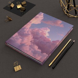 May include: A rectangular hardback notebook featuring a printed cover design of pink and purple clouds against a twilight sky with scattered stars. The notebook is positioned alongside two pencils, one gold and one black, and several gold binder clips.