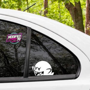 Peut inclure: Un autocollant en vinyle blanc représentant un fantôme de dessin animé avec une expression malicieuse et des dents pointues sur une vitre de voiture, accompagné d'un autocollant circulaire avec le texte rose et bleu Modified Cars 74.