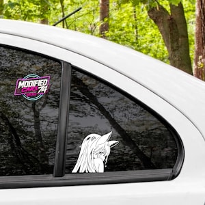 Könnte beinhalten: Ein weißer Vinyl-Aufkleber auf einer Autoscheibe, der eine stilisierte Anime-Figur mit langem Haar und Tierohren zeigt, neben einem kreisförmigen Logo-Aufkleber mit der Aufschrift MODIFIED CARS 74 in weißer und rosa Schrift mit neonblauer Umrandung.
