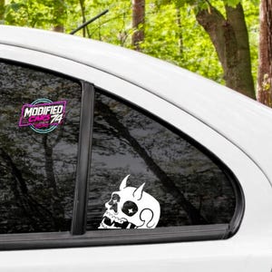 May include: A white vinyl decal of a horned skull with star-shaped eye sockets applied to a car window. Above the skull, a circular logo features the text MODIFIED CARS 74 in pink and white lettering against a dark background.