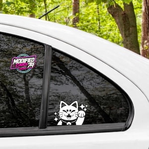 May include: A white vinyl decal of a beckoning cat with sparkles applied to a car window. Above it, a second decal features the text MODIFIED CARS 74 in a stylized pink and cyan circular frame. The decals are placed on the glass of a white vehicle.