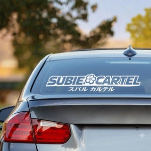 Puede incluir: Una calcomanía de vinilo blanca aplicada en la ventana trasera de un coche gris oscuro. La calcomanía presenta el texto SUBIE CARTEL en una fuente estilizada en negrita con un icono de flor de cerezo en el centro y el texto japonés スバル カルテル debajo.