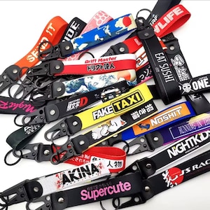 Peut inclure: Plusieurs porte-clés avec des mousquetons en métal noir et des lanières en tissu coloré. Les lanières présentent du texte japonais, des graphiques et des phrases comme "FAKE TAXI", "EAT SUSHI" et "Supercute". Les couleurs incluent le rouge, le jaune, le bleu et le noir.