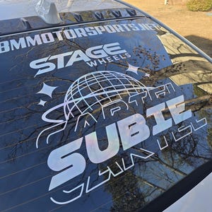 Może przedstawiać: Tylna szyba białego samochodu prezentuje dużą naklejkę z napisami "BMMOTORSPORTS.NET", "STAGE WHEELS" i "SUBIE". Projekt zawiera globus i gwiazdy, wszystko w odblaskowym, iryzującym wykończeniu.
