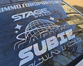 Könnte beinhalten: Die Heckscheibe eines weißen Autos zeigt einen großen Aufkleber mit den Texten "BMMOTORSPORTS.NET", "STAGE WHEELS" und "SUBIE". Das Design umfasst eine Weltkugel und Sterne, alles in einem reflektierenden, irisierenden Finish.