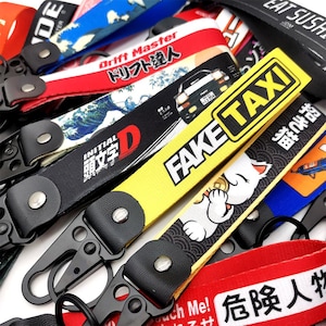 May include: A collection of colorful keychains with various designs and text. Keychains include the words "FAKE TAXI", "INITIAL D", and "Drift Master". The keychains have black metal clasps and leather accents.