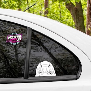 Puede incluir: Una calcomanía de vinilo blanco aplicada en la ventana de un coche con una ilustración de líneas de un personaje de anime con cuernos, mostrando solo los ojos y las manos haciendo un gesto, junto a un logo estilo neón MODIFIED CARS 74.