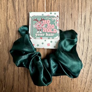 Puede incluir: Un scrunchie de satén verde con una tarjeta navideña festiva que dice "Ho Ho Hold your hair".