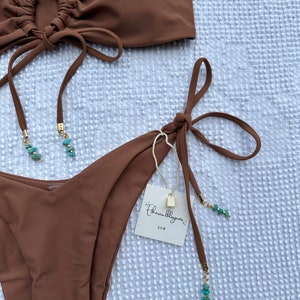 May include: A brown bandeau bikini top with a gathered detail and a tie closure. The matching bikini bottom has a high-cut leg and a tie closure on each side. The bikini is adorned with turquoise beads.