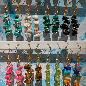 Puede incluir: Una muestra de 12 pares de pendientes de piedras preciosas envueltos en oro. Cada par presenta un tipo diferente de piedra preciosa, incluyendo turquesa, blanco, verde, negro, rosa, ojo de tigre, y más.