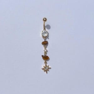 Puede incluir: Un piercing para el ombligo de color dorado con una gema clara y redonda en la parte superior y un dije de estrella en la parte inferior. El anillo presenta una serie de pequeñas piedras marrones de forma irregular y gemas claras suspendidas.