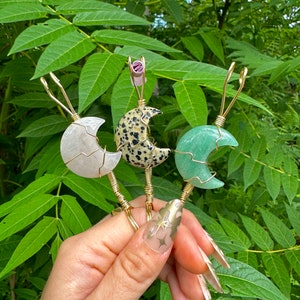 May include: Three crescent moon-shaped crystal pendants. The pendants are made of white, speckled, and green materials. Each moon is wrapped in gold wire and has a gold wire loop. The background is green foliage. The pendants are held in a hand.