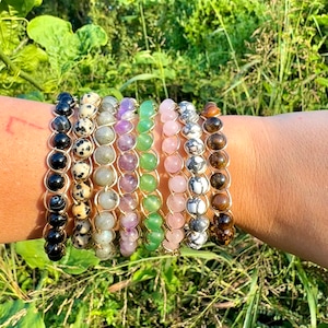 May include: A stack of eight bracelets made with different colored gemstones, including black, brown, white, green, purple, and pink. The bracelets are strung on gold chains.