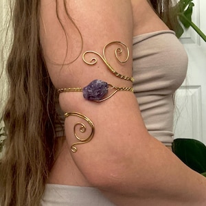 May include: Gold-toned arm cuff featuring a raw amethyst stone. The cuff has a delicate, swirling design that wraps around the arm. The amethyst is a deep purple color, adding a touch of natural beauty to the jewelry.
