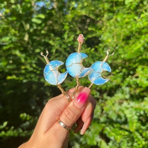 May include: Three gold wire wrapped crescent moon shaped opalite crystal charms. The charms are held in a hand with pink nail polish.