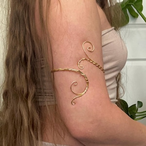 May include: A gold-colored arm cuff with a delicate, swirling design. The cuff wraps around the upper arm and features intricate, curved wire detailing. The jewelry piece is a stylish accessory.
