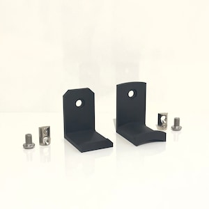 May include: Two black metal wall brackets with rounded edges and a single hole in the centre. The brackets are designed to hold a shelf or other object. The image also shows four silver screws and two silver metal brackets.