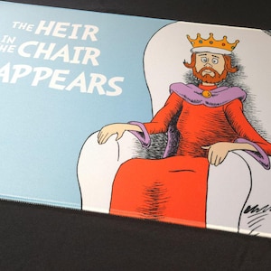 May include: A mousepad featuring a cartoon illustration of a king in a throne. The king wears a crown and red robe. The text "THE HEIR IN THE CHAIR REAPPEARS" is printed in white on a light blue background.