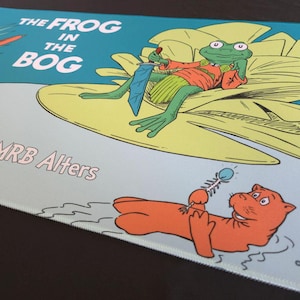 May include: A mousepad featuring a whimsical illustration from "The Frog in the Bog" by MRB Alters. The design includes a frog in a red shirt and a cartoon otter. The text "THE FROG IN THE BOG" is displayed at the top.