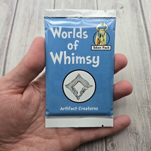 Puede incluir: Un paquete de fichas azul titulado "Worlds of Whimsy" con las palabras en blanco. El paquete presenta un gráfico circular de una criatura y las palabras "Artifact Creatures". También es visible un pequeño gráfico que dice "Token Pack".