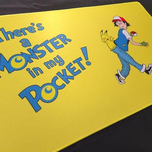 May include: Yellow mousepad with the text "There's a MONSTER in my POCKET!" in blue. A cartoon illustration of a character with a yellow creature in their pocket is on the right. The mousepad has a smooth surface.
