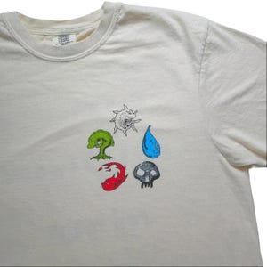 May include: An off-white short-sleeve t-shirt featuring a circular arrangement of five small, colorful graphic illustrations. The designs include a green tree-like creature, a white sun-like shape, a blue water drop, a red flame, and a gray figure.