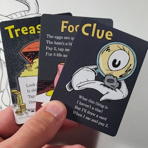 May include: Three black cards with white text and illustrations. The first card says "Treasure" and has a cartoon image of a treasure chest with gold coins. The second card says "Food Clue" and has a cartoon image of a plate of eggs and ham. The third card says "Food Clue" and has a cartoon image of a magnifying glass with a single eye looking at a small rock.