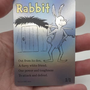 Op de afbeelding: Een verzamelkaart met een cartoonkonijn dat een zwaard vasthoudt. De kaart heeft een blauwe en zilveren achtergrond met de tekst "Rabbit" in gele letters. De tekst op de kaart luidt: "Out from its den, A furry white friend. One power and toughness To attack and defend. &copy;2024 MRB Alterti 1/1"