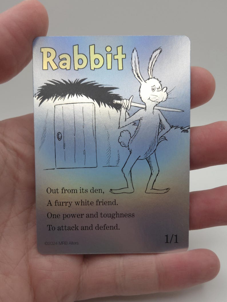 Op de afbeelding: Een verzamelkaart met een cartoonkonijn dat een zwaard vasthoudt. De kaart heeft een blauwe en zilveren achtergrond met de tekst "Rabbit" in gele letters. De tekst op de kaart luidt: "Out from its den, A furry white friend. One power and toughness To attack and defend. &copy;2024 MRB Alterti 1/1"