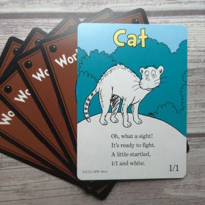 May include: A stack of children's flashcards. The front card features a white cat illustration with the word "Cat" in yellow. The background is blue with green tree shapes. The card includes the text "Oh, what a sight! It's ready to fight. A little startled, 1/1 and white."