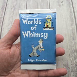 May include: A blue "Worlds of Whimsy" token pack with a wizard illustration. The pack includes the text "Trigger Reminders" and "Token Pack". The packaging is sealed and held in a hand.