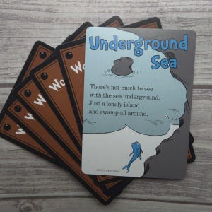 Könnte beinhalten: Ein Stapel Spielkarten mit braunen Rändern und dem Buchstaben "W". Die oberste Karte zeigt den Text "Underground Sea" und eine Illustration eines Felsens, Wassers und eines blauen Fisches. Die Karte enthält auch den Text "There's not much to see with the sea underground. Just a lonely island and swamp all around."
