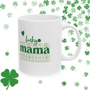 Könnte beinhalten: Weiße Keramiktasse mit einem grünen Kleeblatt und dem Text "lucky mama" in grünen Buchstaben. Der Text wird in kleinerer Schriftgröße mit einer goldenen Umrandung wiederholt.