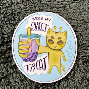 Op de afbeelding: Een sticker met een cartoonkat met zes ogen die een paarse en blauwe boba-thee vasthoudt met de tekst "Need my sweet treat".